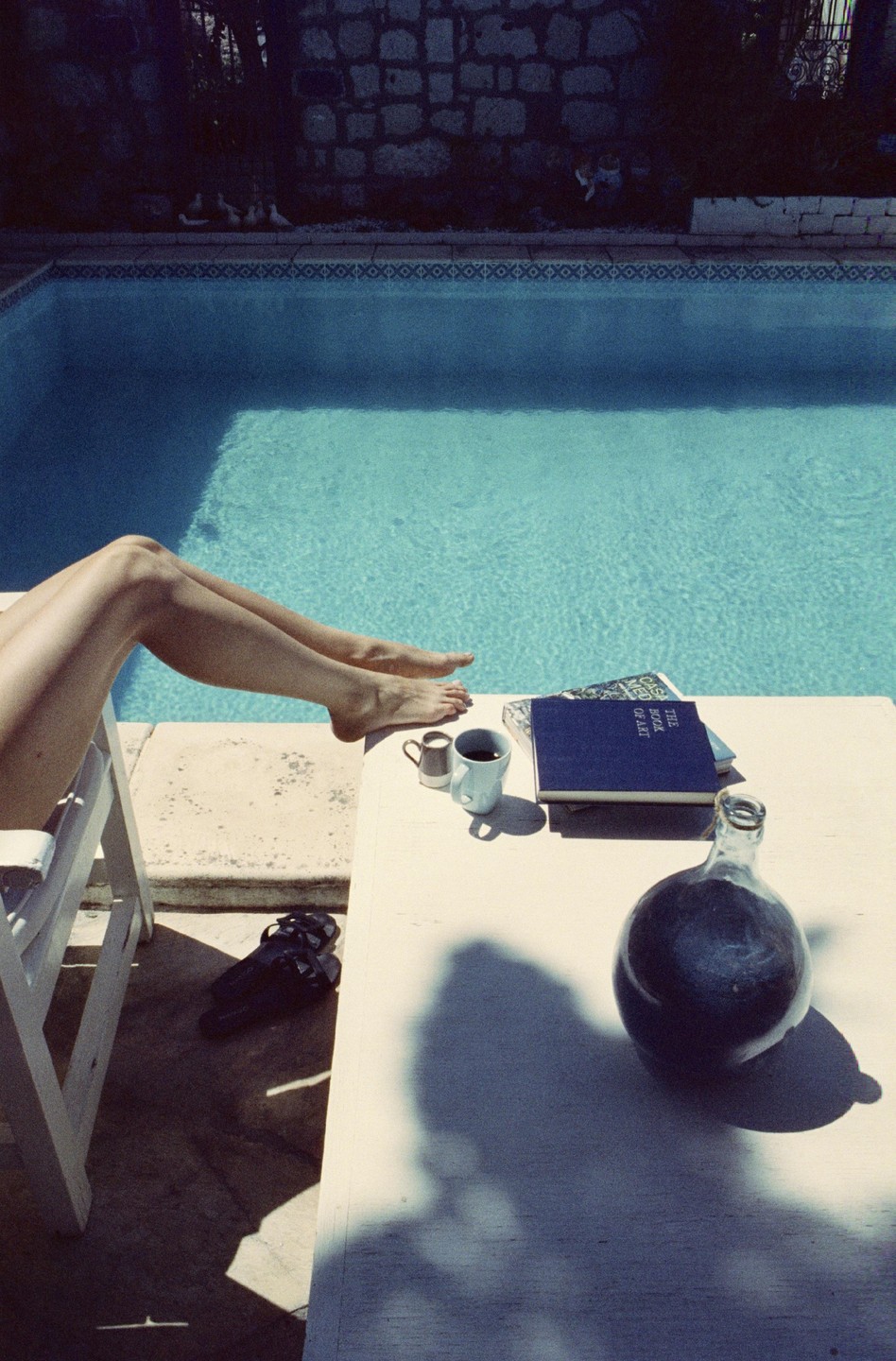 A person's legs are propped up next to a swimming pool with a table holding a book, a cup, and a bottle.