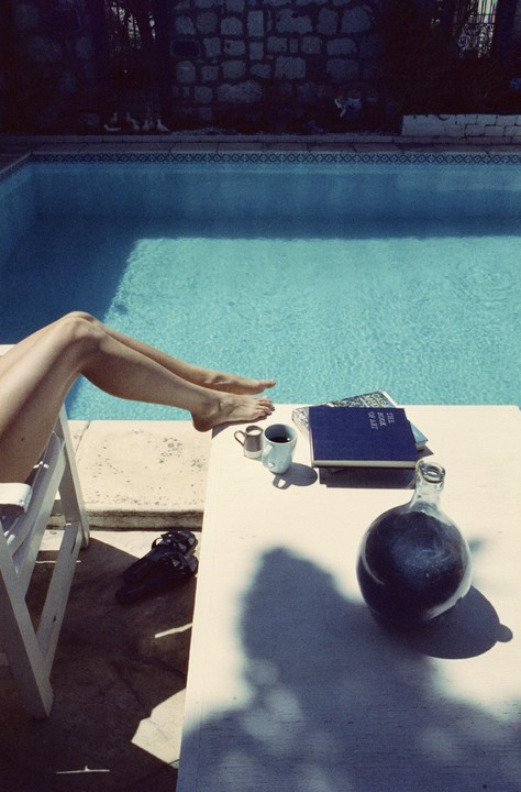 A person's legs are propped up next to a swimming pool with a table holding a book, a cup, and a bottle.