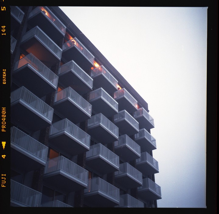 A tall building with many balconies is illuminated by warm lights from within, set against a pale sky.