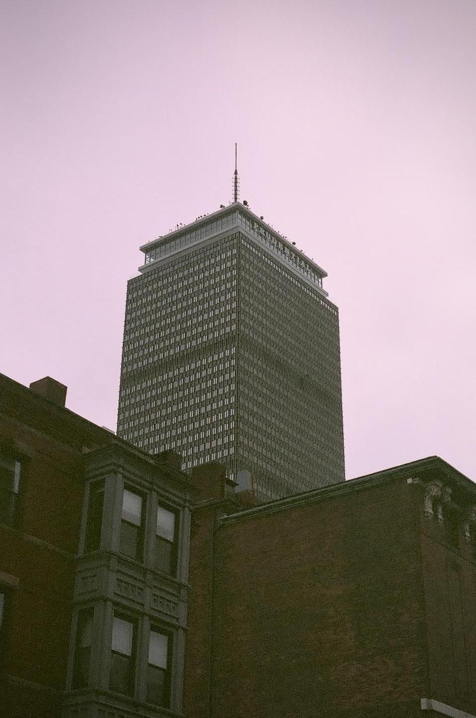 A tall skyscraper with a spire on top rises above brick buildings under a pink sky.