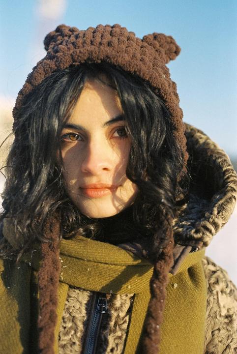 A woman with dark, curly hair wears a brown knitted hat with ears, a green scarf, and a fur-lined coat.