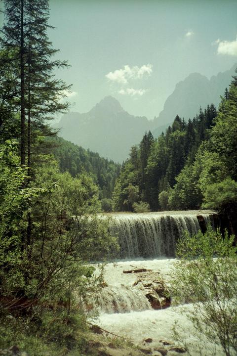 A waterfall cascades over rocks into a river, surrounded by a forest and mountains under a hazy sky.