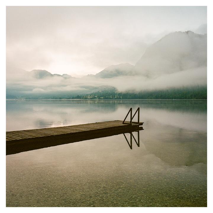 A wooden dock with a ladder extends into a calm lake surrounded by fog-covered mountains.