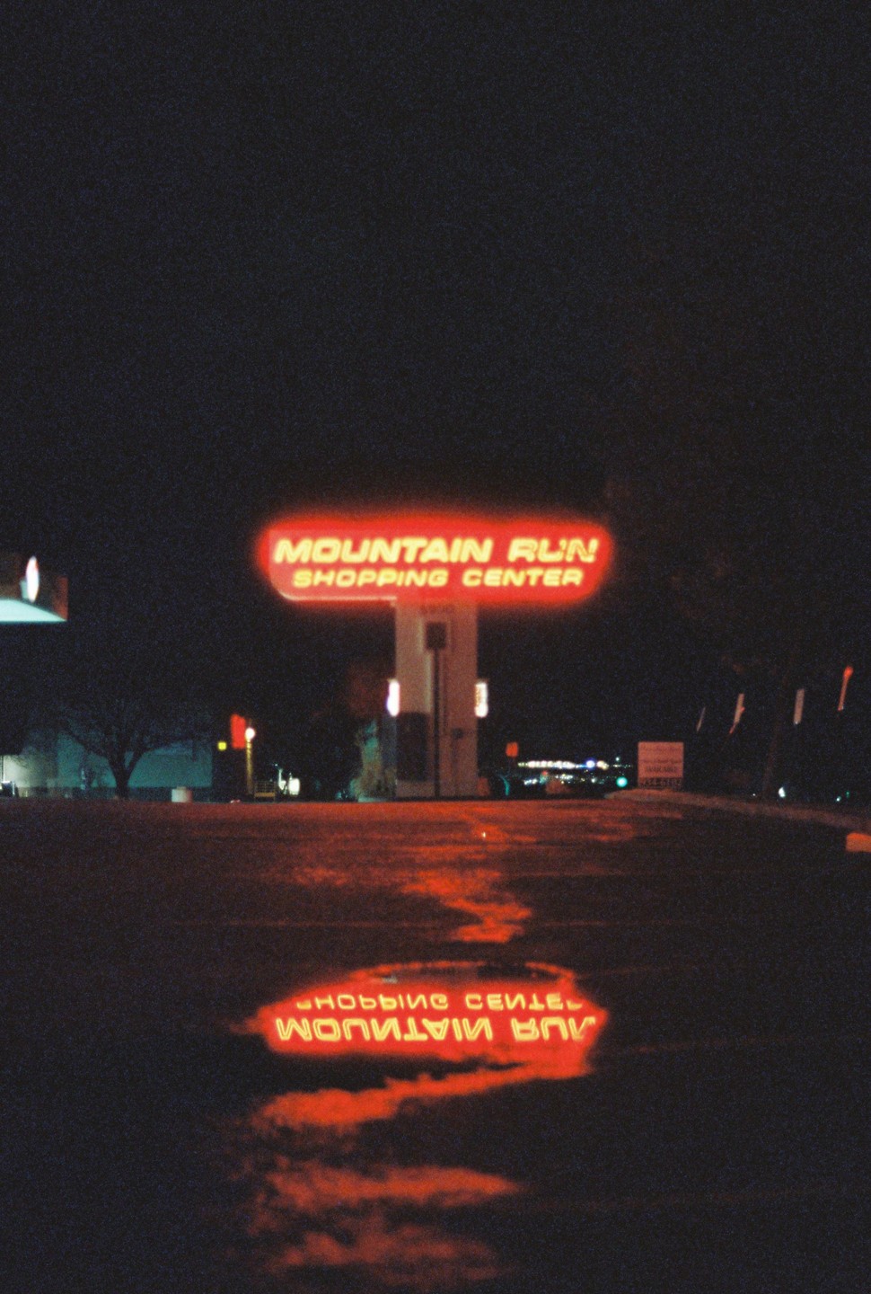 A neon sign for Mountain Run Shopping Center is reflected in a puddle at night.