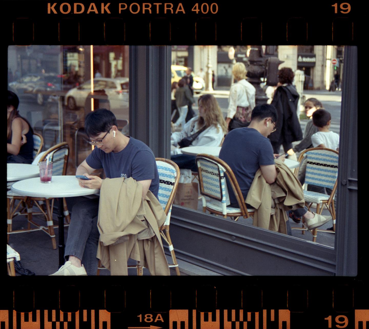 A man sits at an outdoor cafe table looking at his phone, with other people and a street visible in the background.