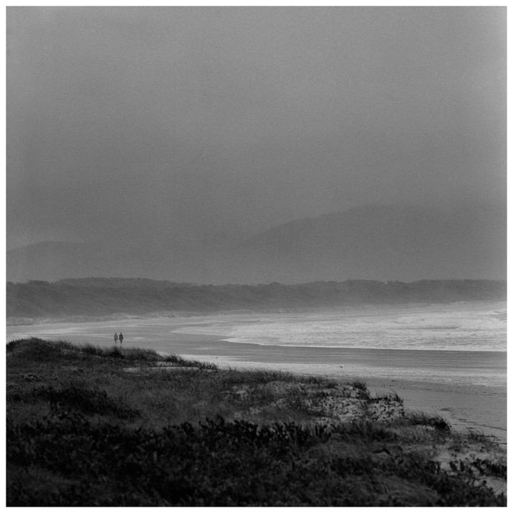 Two people walk along a misty beach with dunes and mountains in the background.