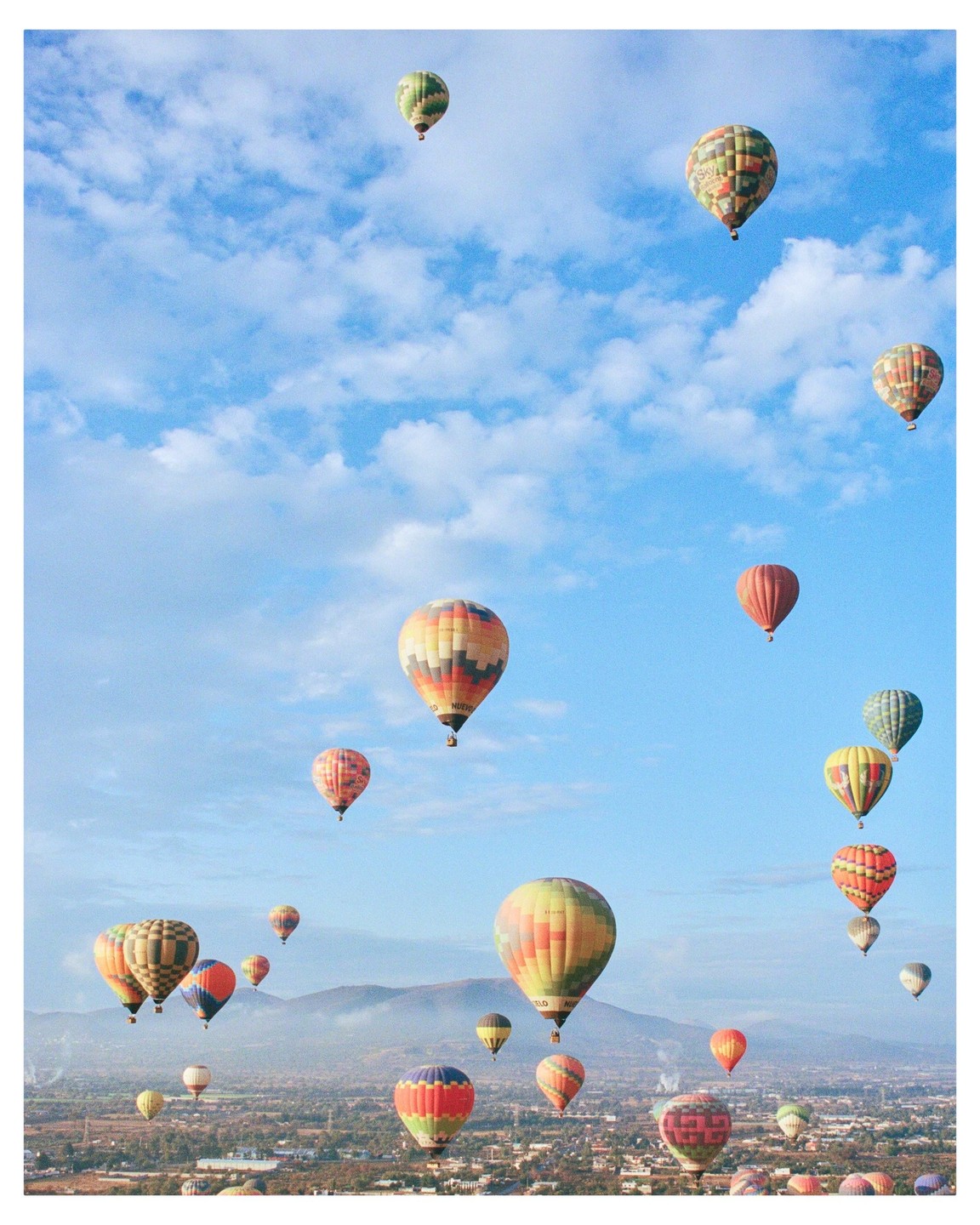 Many hot air balloons float in a blue sky above a town and mountains.
