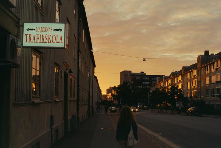 A woman walks down a street with buildings and a sign for a driving school under a golden hour sky.