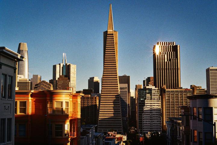 A cityscape with a prominent triangular skyscraper and other buildings is illuminated by golden hour sunlight.