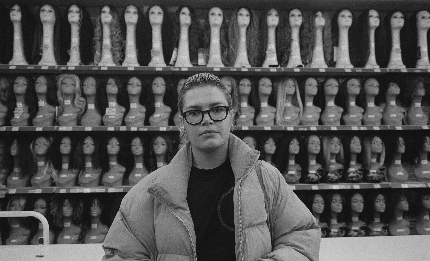 A woman wearing glasses and a puffer jacket stands in front of shelves filled with mannequin heads wearing wigs.