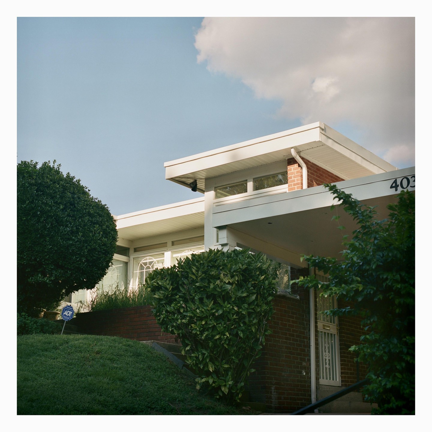 A modern house with a flat roof and brick accents is surrounded by lush green bushes and grass under a partly cloudy sky.