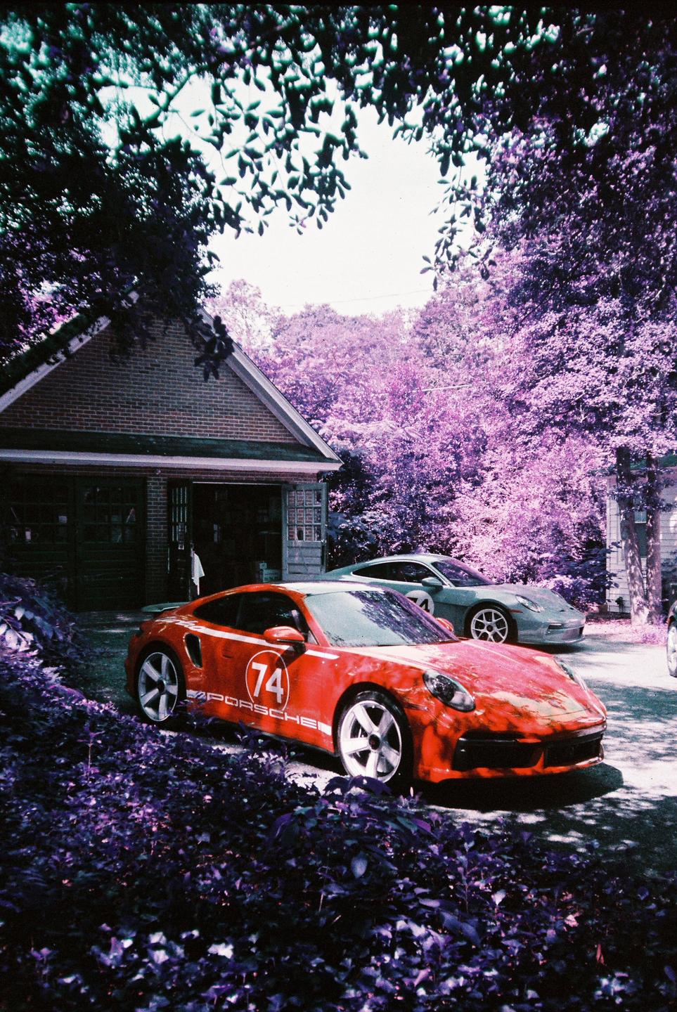 A red sports car with the number 74 on its side is parked in a driveway next to a brick garage, with purple-hued trees in the background.