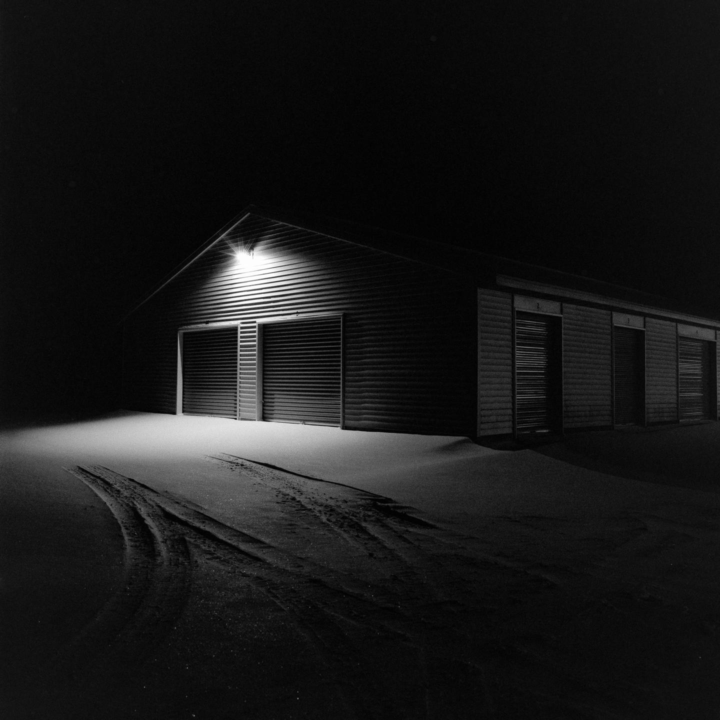 A building with multiple garage doors is illuminated by a single light at night, with snow covering the ground and tire tracks visible.