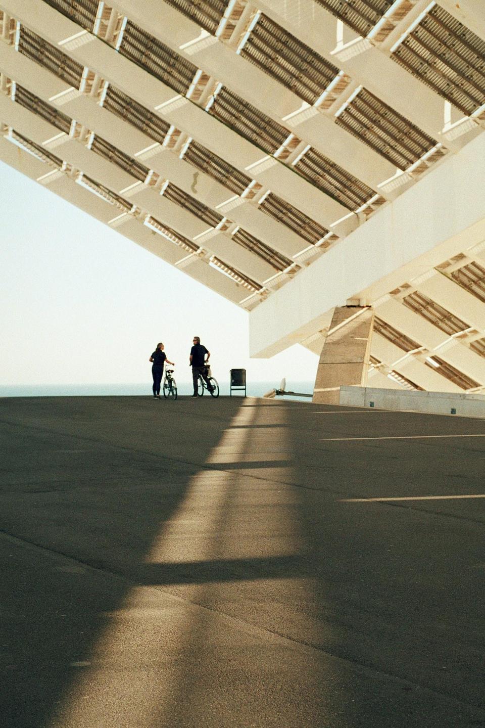 A couple stands with their bicycles on an asphalt surface under a large, modern architectural structure with the ocean visible in the distance.