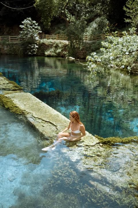 A woman with red hair sits in clear blue water next to a mossy stone structure, surrounded by lush green foliage.