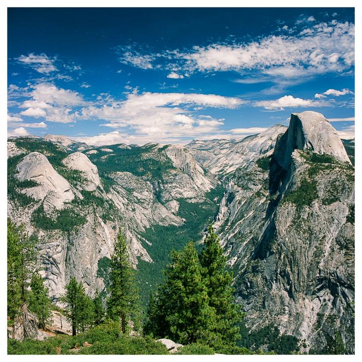 A wide view of Yosemite Valley shows Half Dome and other granite formations under a blue sky with white clouds.