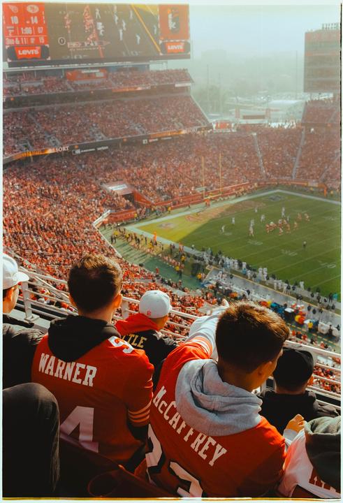 A crowd of people watches a football game from the stands of a stadium.