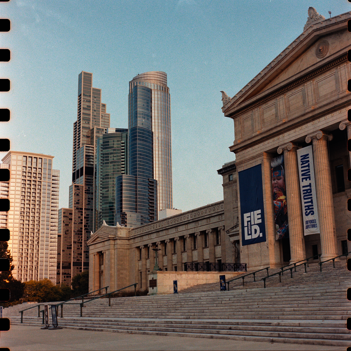 A grand museum building with large steps and banners stands in front of modern skyscrapers under a clear sky.