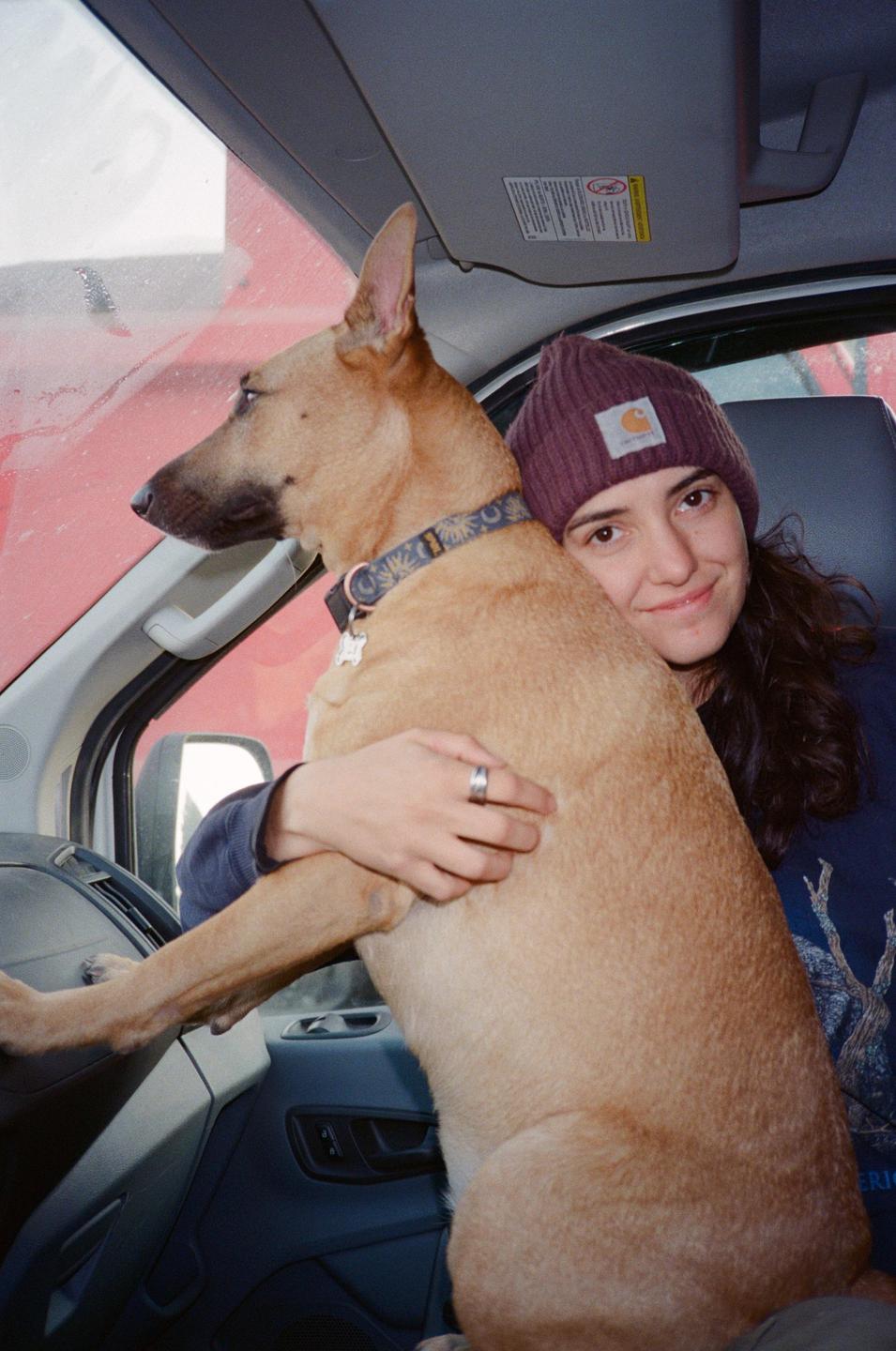 A woman wearing a beanie holds a dog in the driver's seat of a vehicle.