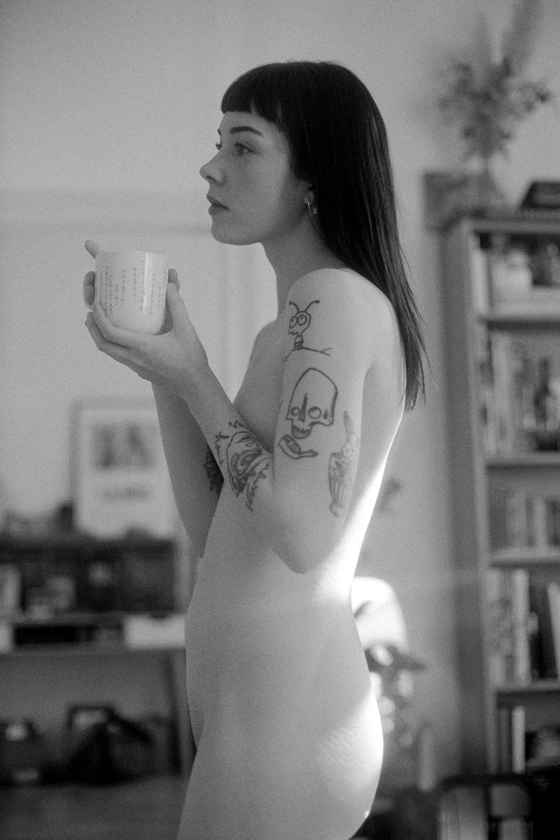 A woman with tattoos on her arms holds a mug and looks to the side.