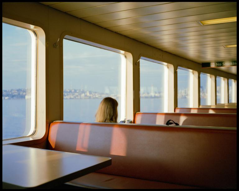 A woman sits on a ferry looking out at a city across the water during golden hour.