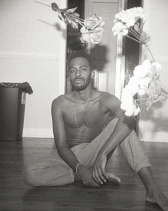 A man sits on a wooden floor surrounded by flowers.
