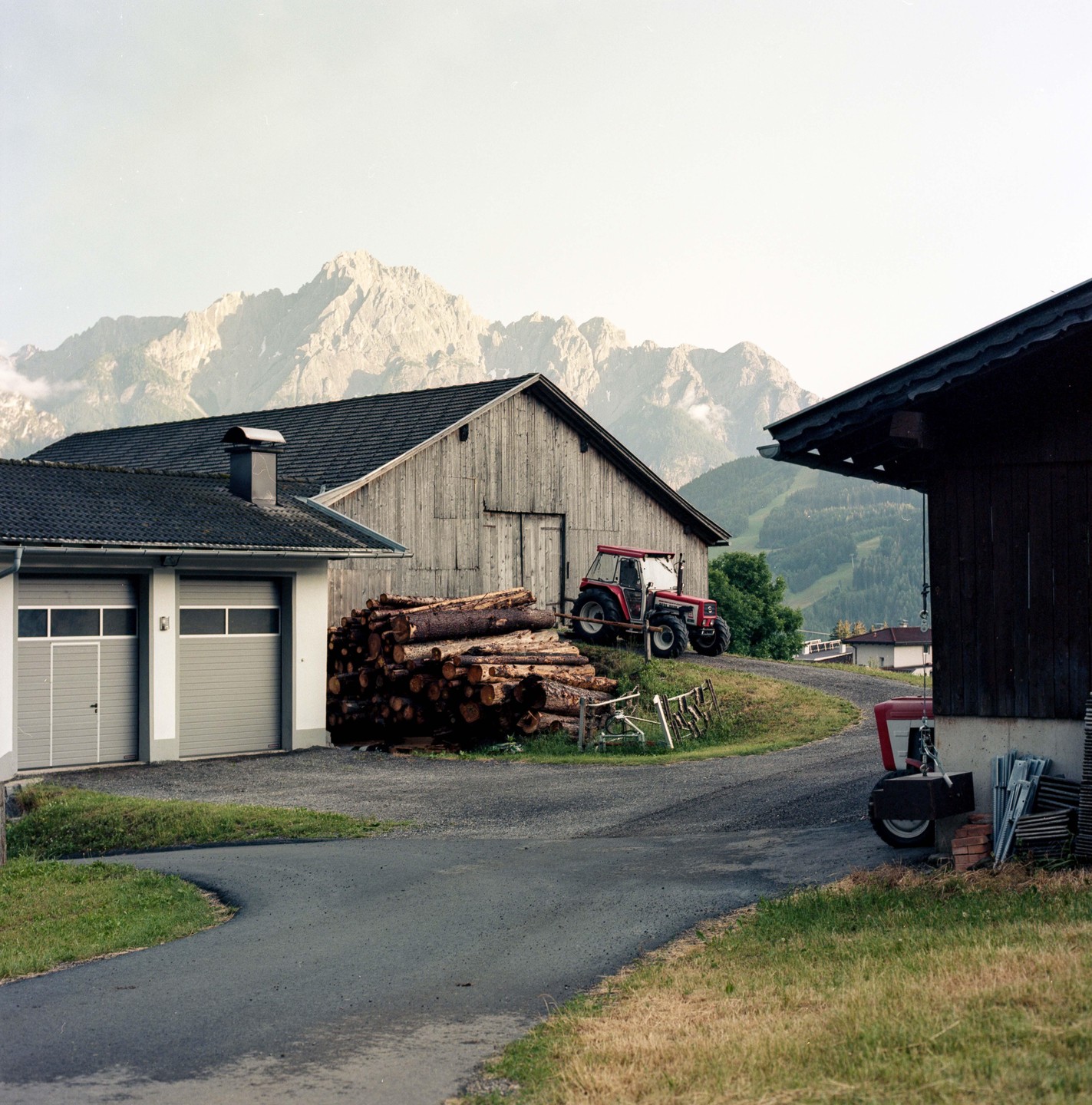 A red tractor is parked on a grassy hill next to a pile of logs and a wooden barn, with mountains in the background.