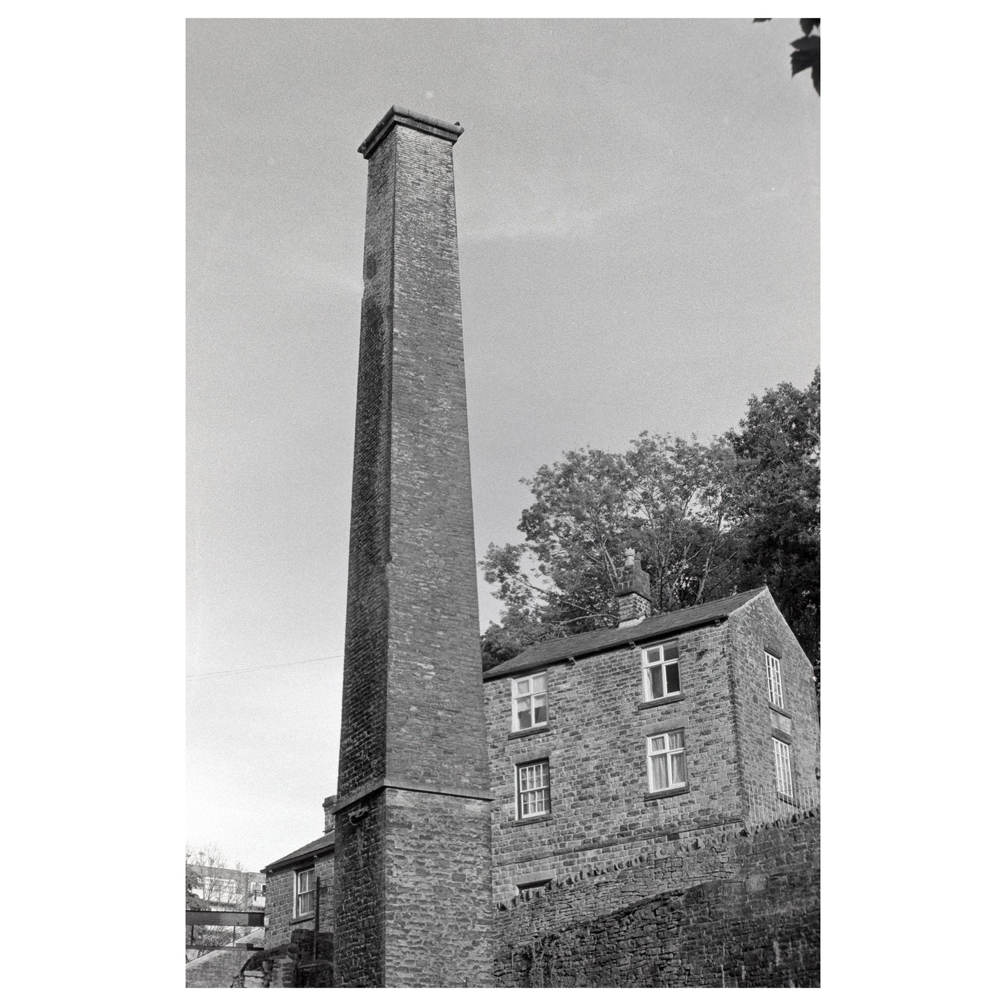 A tall brick chimney stands next to a stone building with trees in the background.