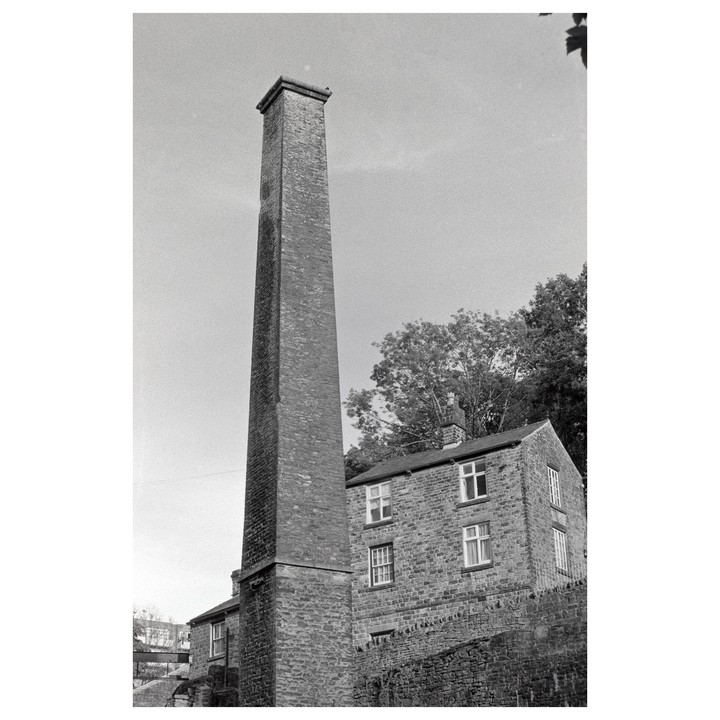 A tall brick chimney stands next to a stone building with trees in the background.
