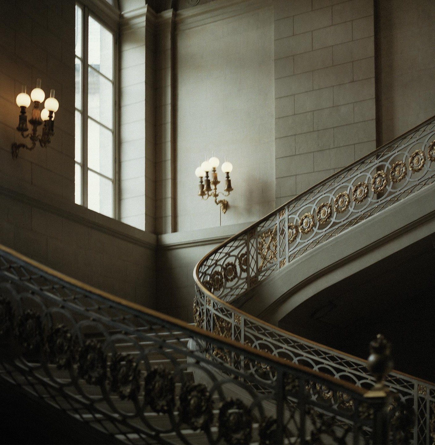 A grand staircase with ornate railings and wall sconces is illuminated by natural light from a large window.