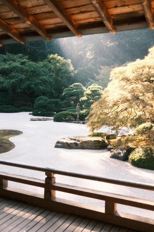 A zen garden with raked sand, rocks, and trees is viewed from under a wooden roof and balcony.