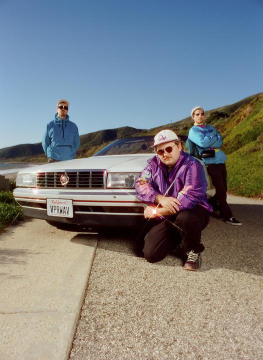 Three men pose with a white car on a coastal road with a hillside and ocean in the background.