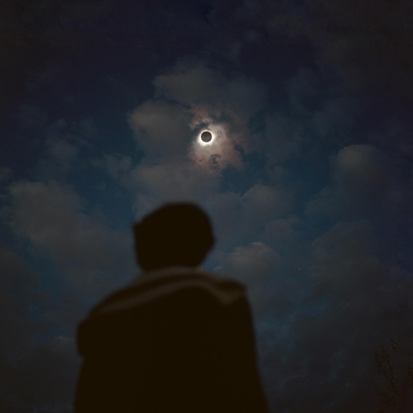A person looks up at a solar eclipse in a cloudy sky.