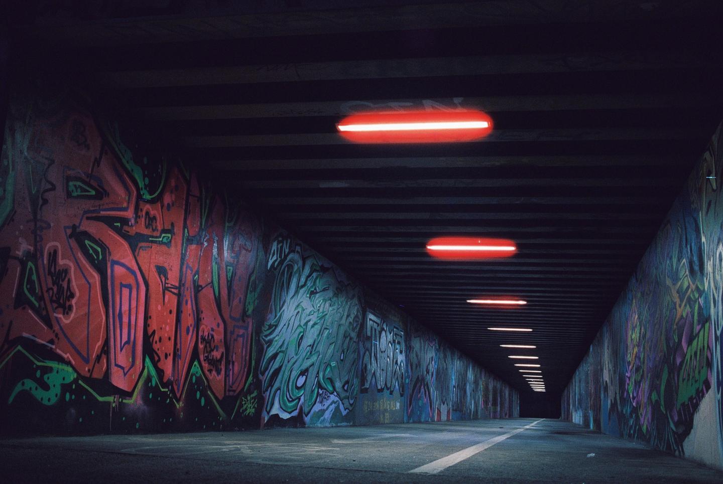 A long, dark underpass is illuminated by a series of red neon lights on the ceiling and covered in colorful graffiti on both walls.