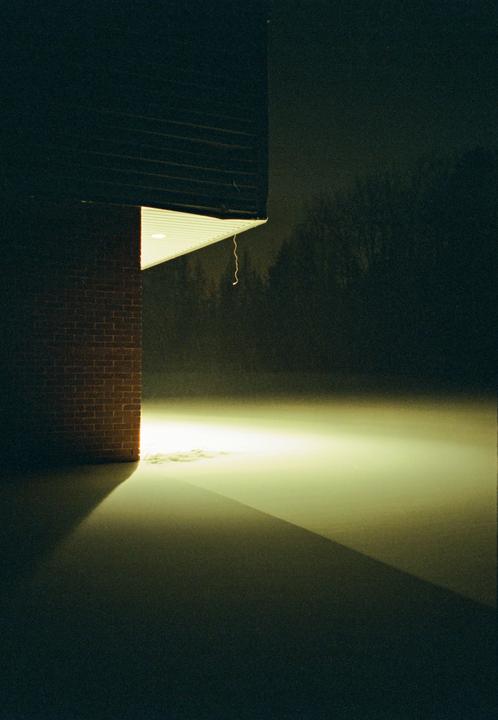 A brick wall and an illuminated overhang cast a shadow on a snow-covered ground at night.