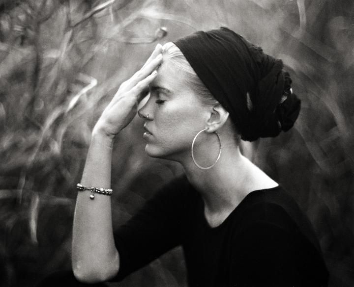 A woman with a headscarf and large hoop earring holds her hand to her forehead with her eyes closed.