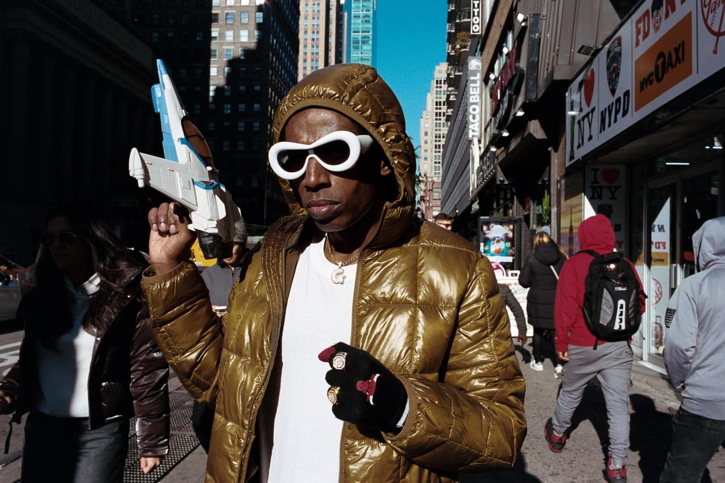 A man in a shiny gold puffer jacket and white sunglasses holds a toy spaceship on a city street.