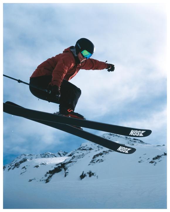 A skier in a red jacket and black helmet is captured mid-air against a backdrop of snow-covered mountains and a cloudy sky.