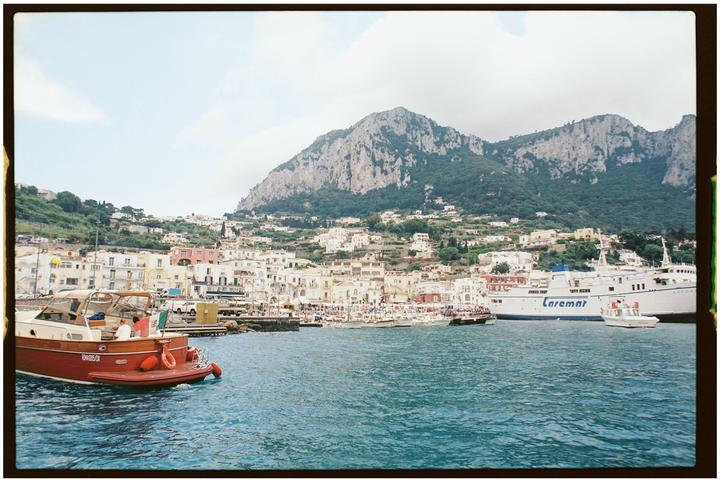 A harbor with boats and buildings is nestled at the base of a large, green mountain.