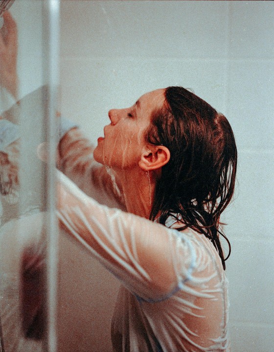 A woman with wet hair and a wet shirt stands in a shower with water running down her face.