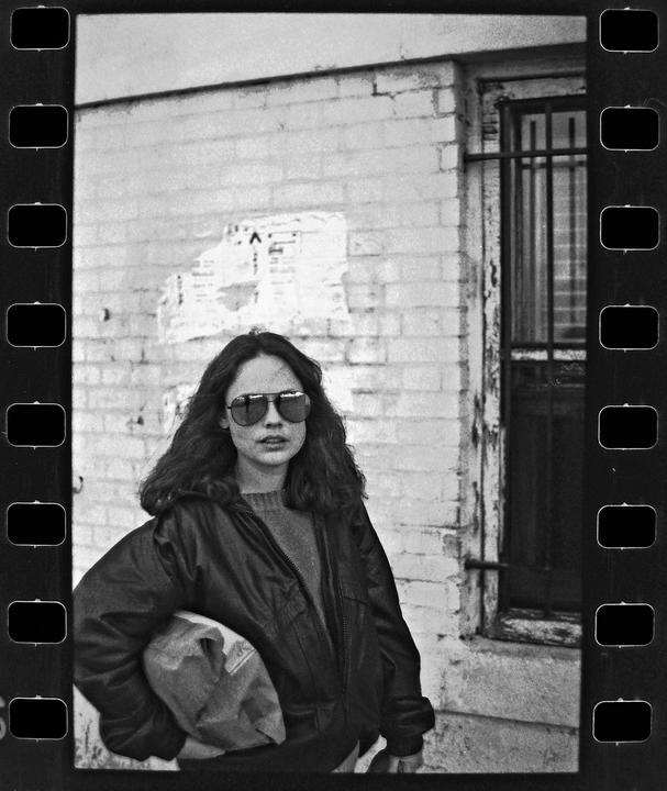 A woman wearing sunglasses and a jacket stands in front of a brick wall with a barred window.