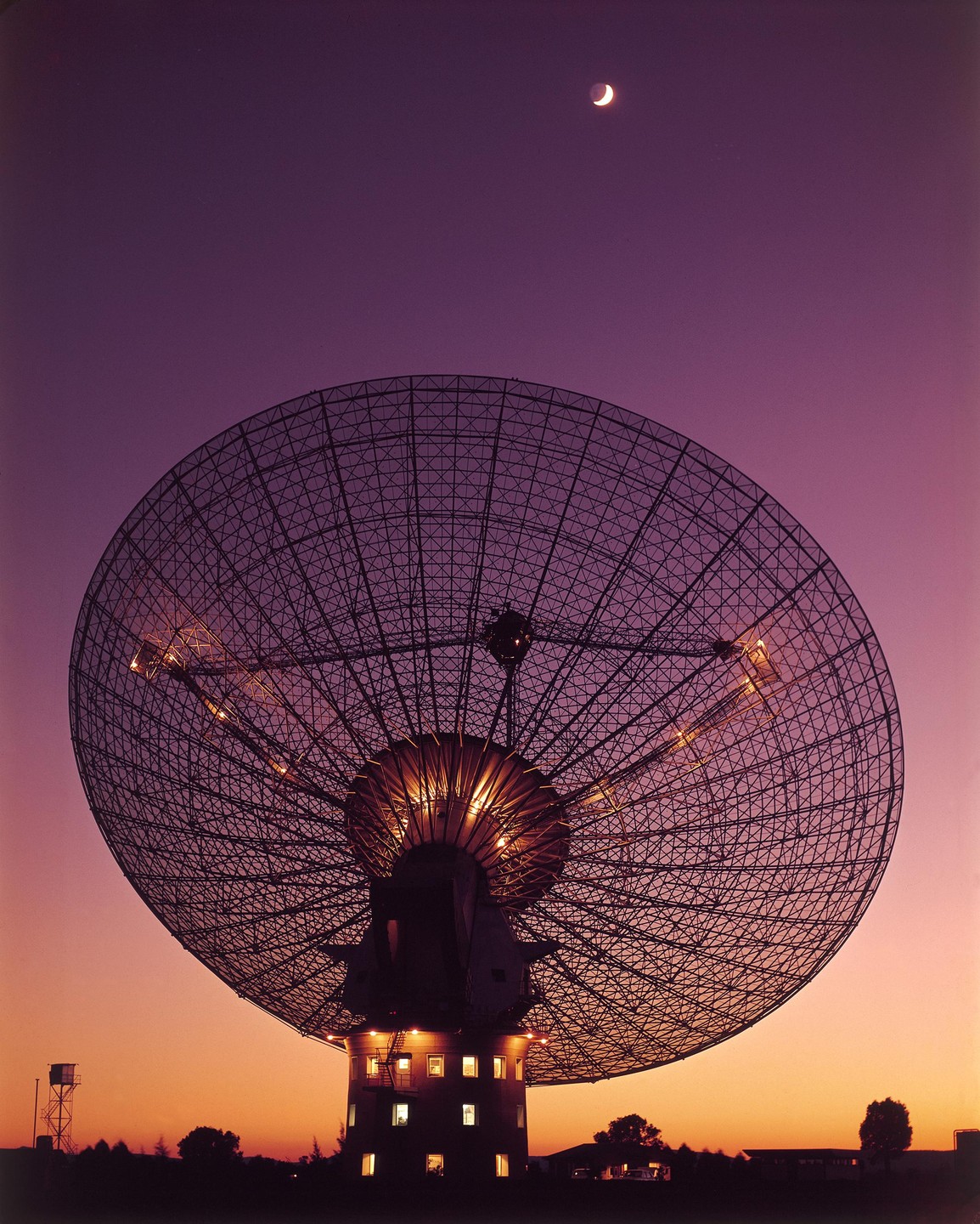 A large radio telescope dish is silhouetted against a vibrant purple and orange sky with a crescent moon.