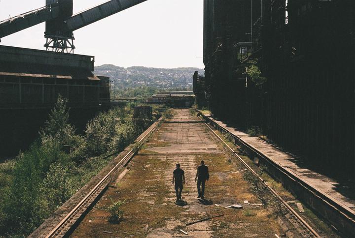 Two men walk away from the camera on an overgrown concrete path between abandoned industrial buildings.