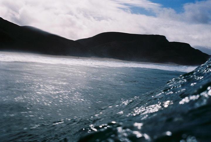 A low-angle view from the ocean shows waves breaking on a beach with dark mountains in the background under a cloudy sky.