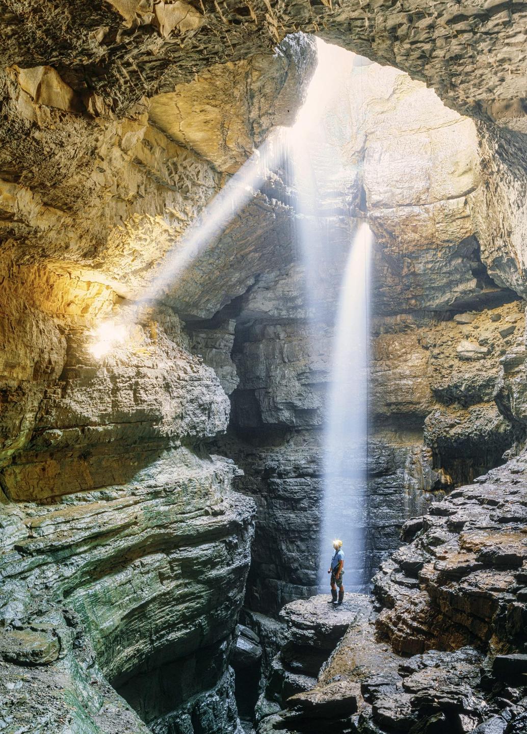A person stands on a rock formation inside a cave with light beams and a waterfall.