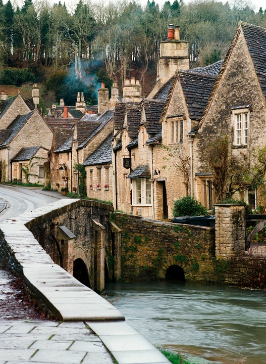 A row of old stone houses with dark roofs lines a street next to a river with a stone bridge.