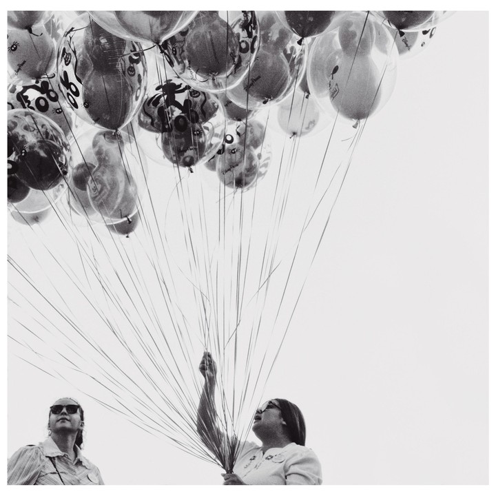 Two women look up at a large bunch of balloons with cartoon characters on them, held by one of the women.