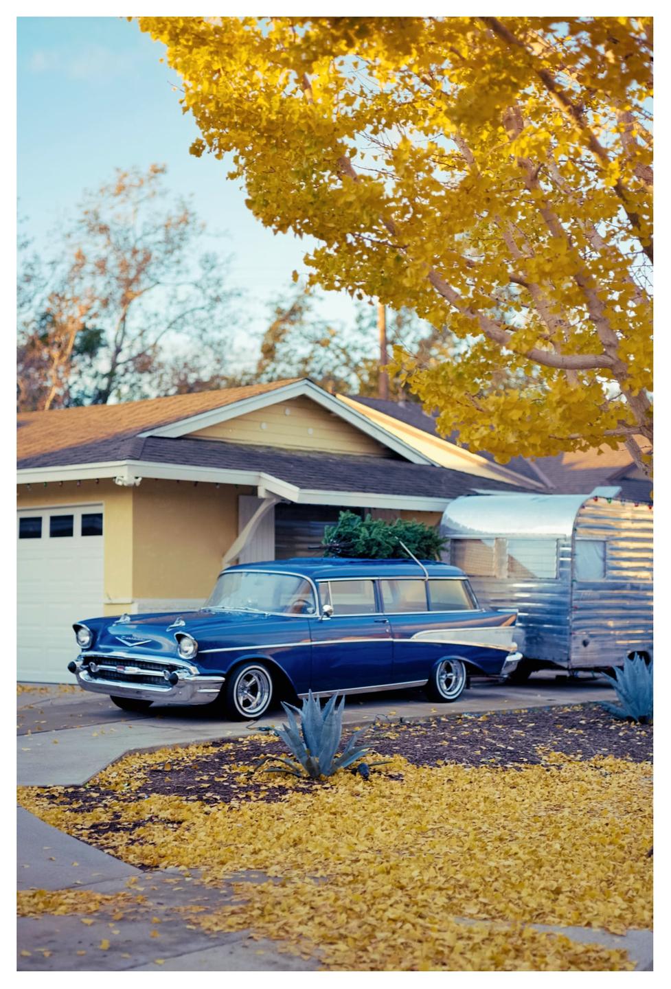 A blue vintage car with a Christmas tree on its roof is parked in a driveway next to a silver trailer, with yellow autumn leaves scattered on the ground.