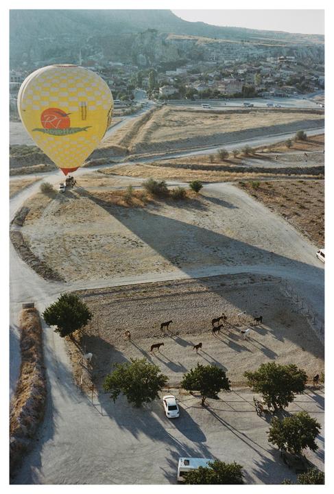 A yellow hot air balloon casts a long shadow over a dry landscape with horses and a distant town.