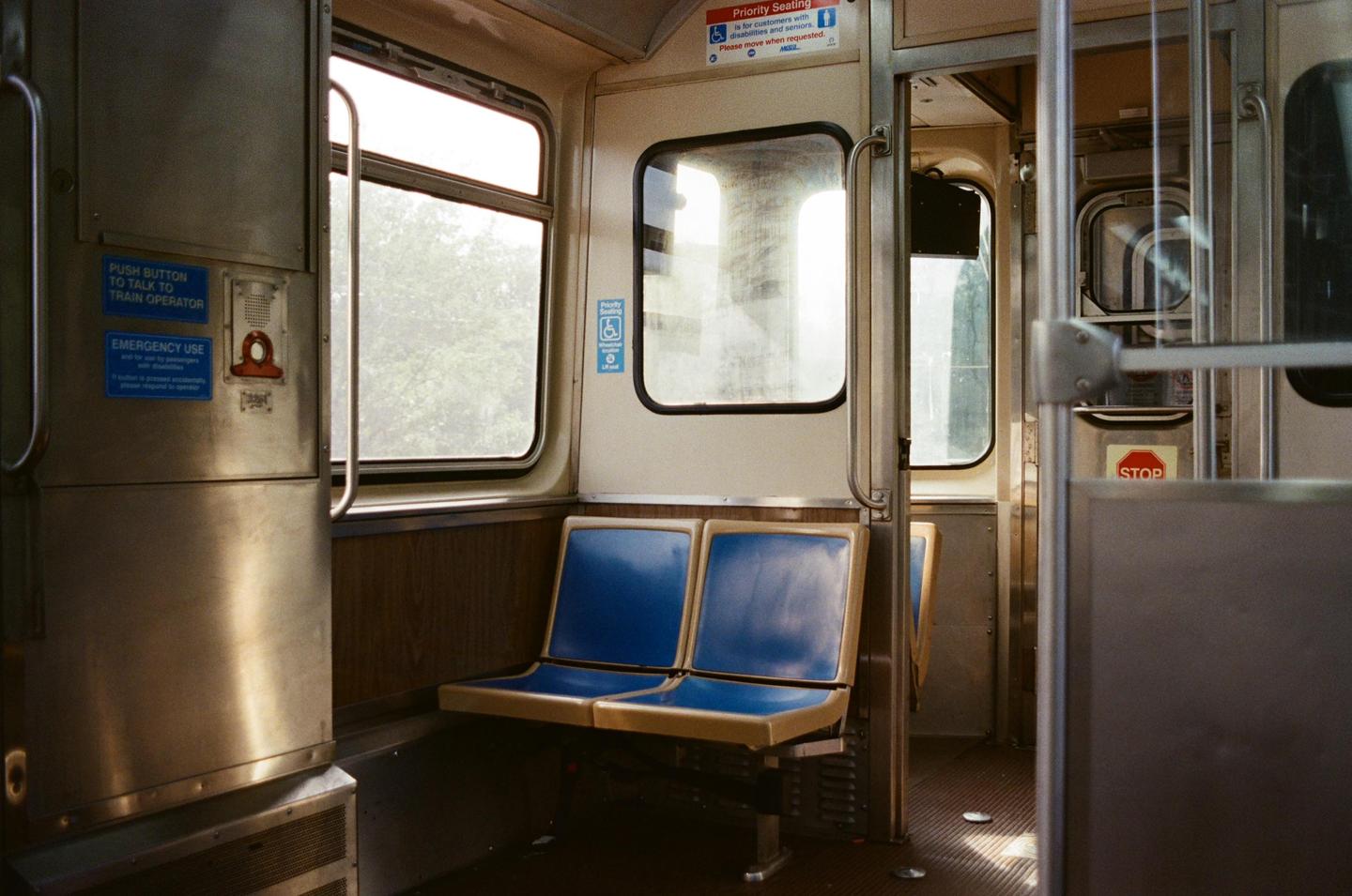 The interior of an empty train car shows blue seats and windows looking out onto green trees.